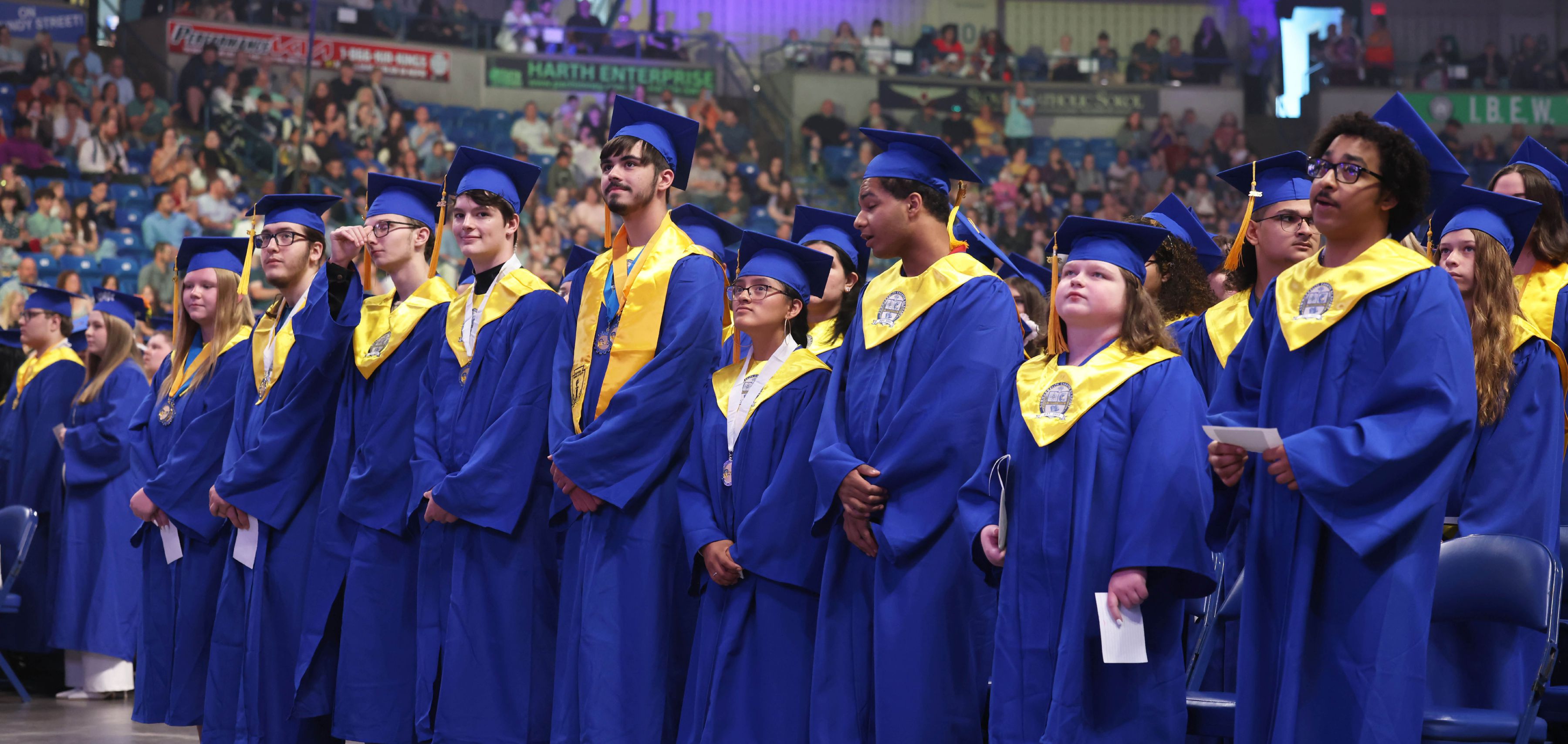 CCA Graduates in blue and yellow caps and gowns standing in a line at a graduation ceremony.